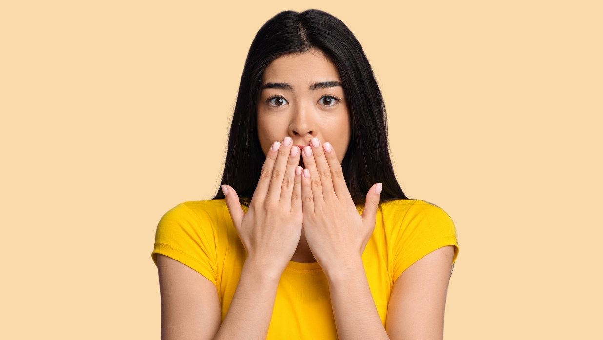 Young woman covering her mouth with both hands, looking surprised or shocked, on a beige background — illustrating the reaction to hearing or saying a swear word.