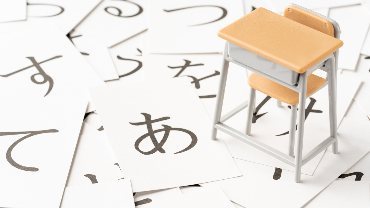 Miniature school desk and chair placed on scattered flashcards featuring basic Japanese hiragana characters.