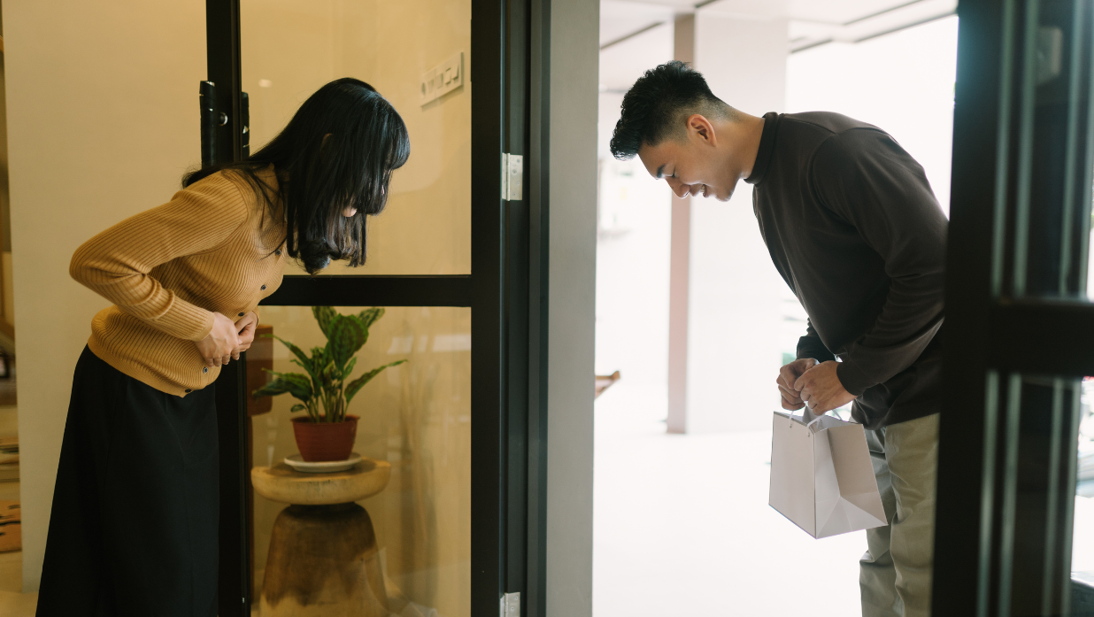 Man and woman bowing respectfully to each other at a doorway, symbolizing Japanese politeness and the cultural nuance behind the phrase 'よろしくお願いします' (yoroshiku onegaishimasu).