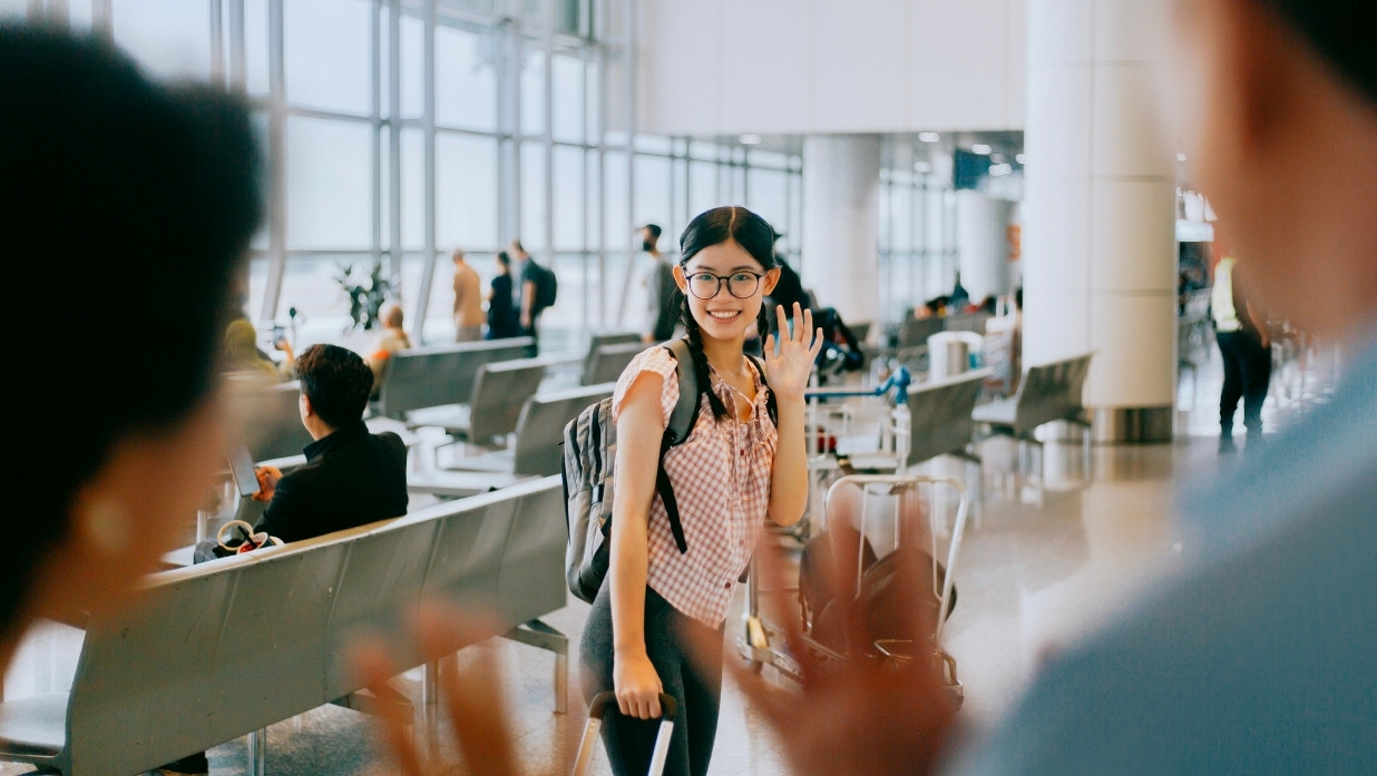 A young woman waves goodbye at an airport terminal, smiling with a suitcase and backpack — symbolizing a farewell scene where someone might say “気をつけて (Ki o Tsukete)” or “Take care” in Japanese.
