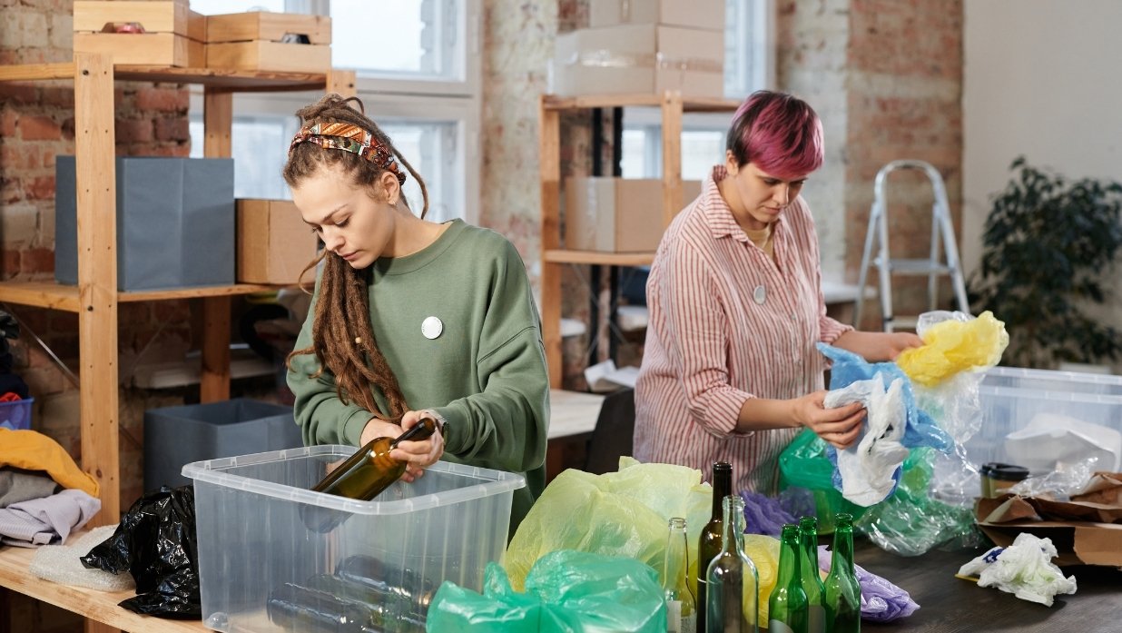 2 people sorting garbage to make sure everything is where it is supposed to be. Plastic bags, glass bottles, and more are on the table. Containers there for organization.