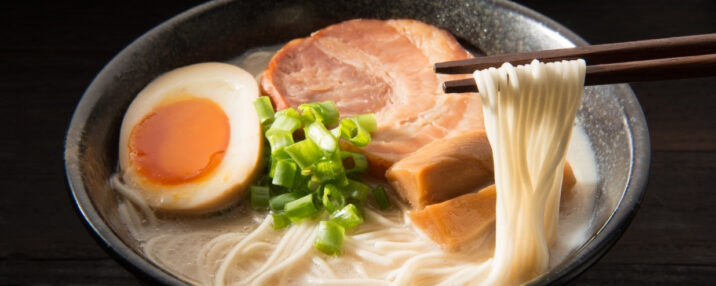 A bowl of ramen with pork, egg, and green onion in it. Noodles being held by chopsticks.