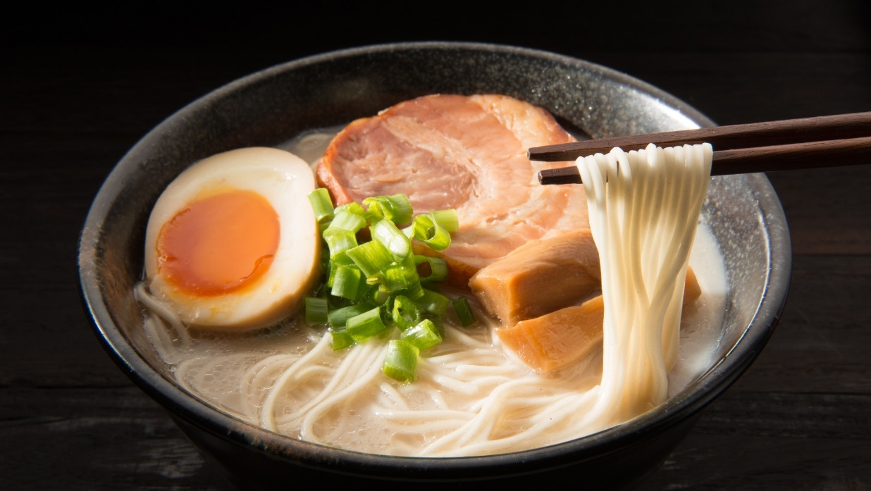 A bowl of ramen with pork, egg, and green onion in it. Noodles being held by chopsticks.