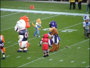 A group of colorful sports mascots standing on an American football field, preparing for an event or show, with a football on the ground.