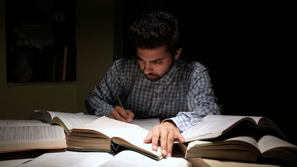 Student surrounded by textbooks studying at a desk. In the dark.