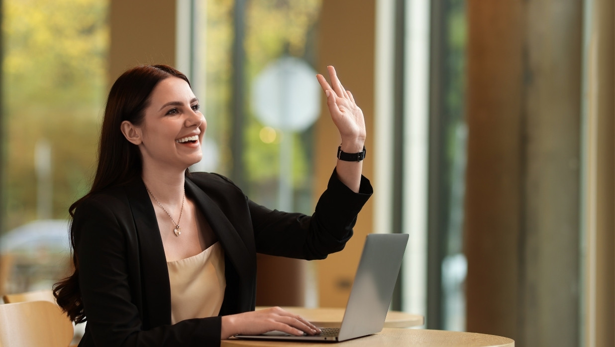 A woman in business clothes waving hello while working at her computer.