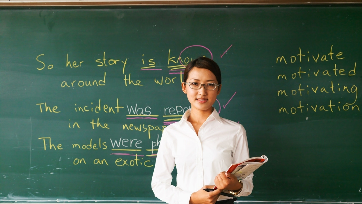 Female teacher teaching a lesson on grammar to class. Chalkboard behind her.