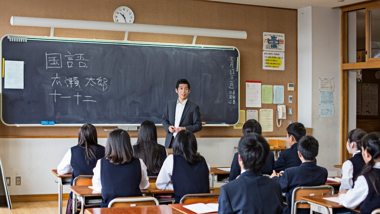 A male teacher at the head of the class teaching his students how to speak/write Japanese.